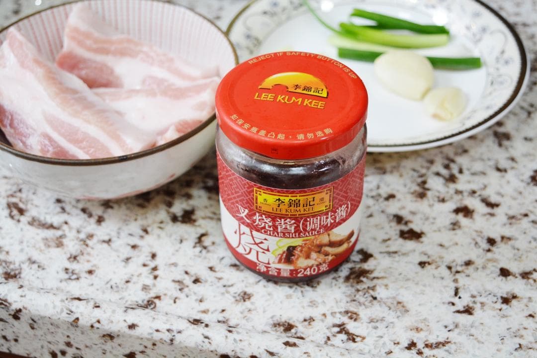 A Chinese-label Lee Kum Kee char siu sauce jar stands in front of raw pork and aromatics.