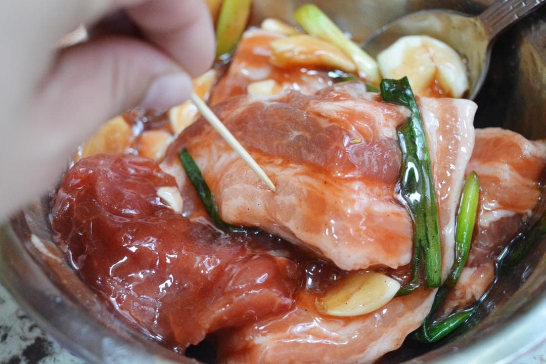 Raw pork pieces coated with red marinade and aromatics, with a hand holding a toothpick over the meat.