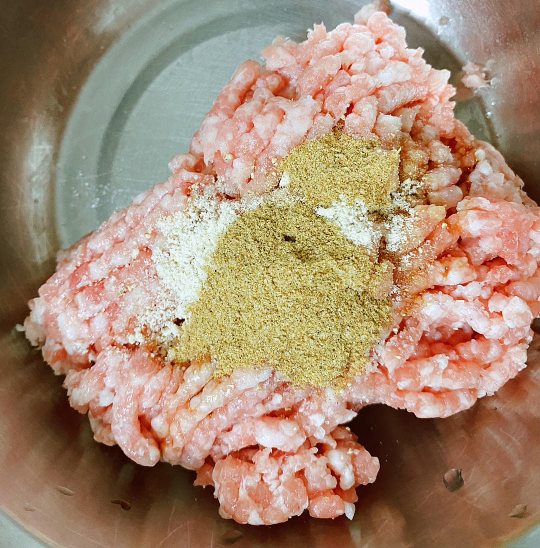 Raw ground pork topped with dry seasonings in a metal mixing bowl.