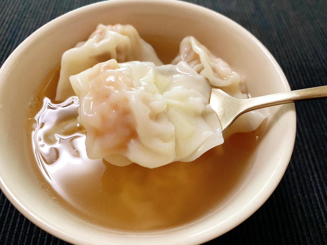 A cooked wonton lifted on a spoon over a bowl of clear brown soup with more wontons.