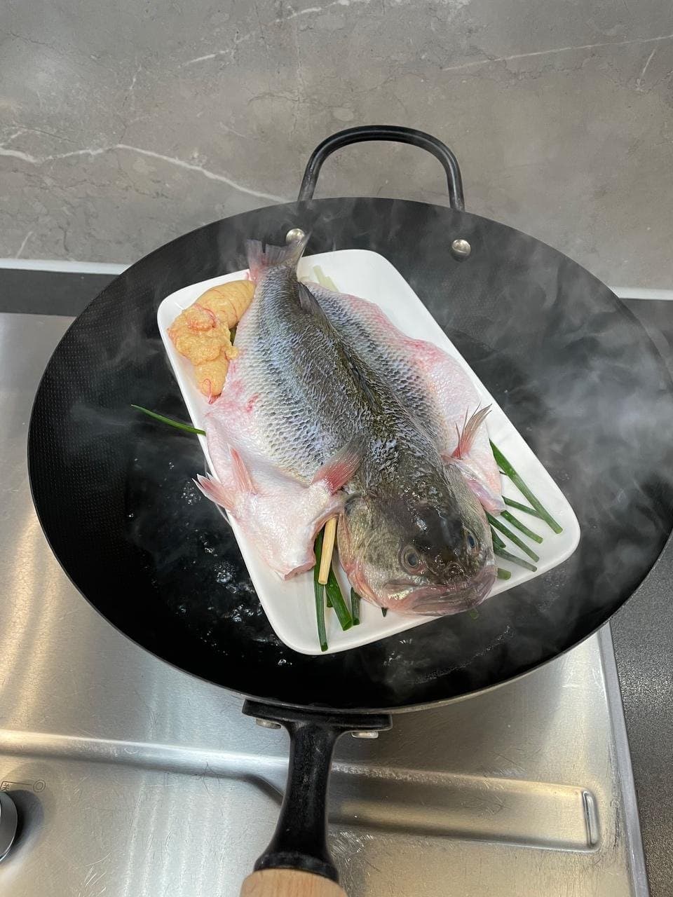 The plate of fish set over steaming water in a wok before the lid is closed.