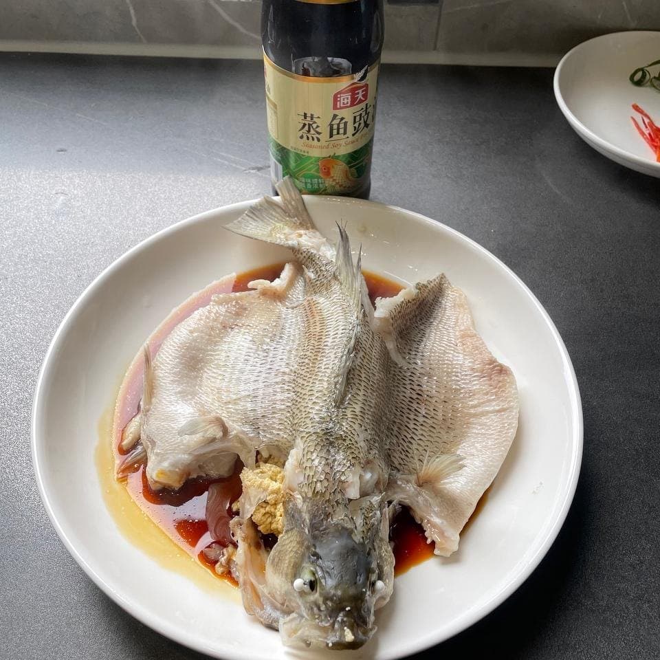 Steamed fish on a clean serving plate with seasoned soy sauce around it, before the fresh garnish.